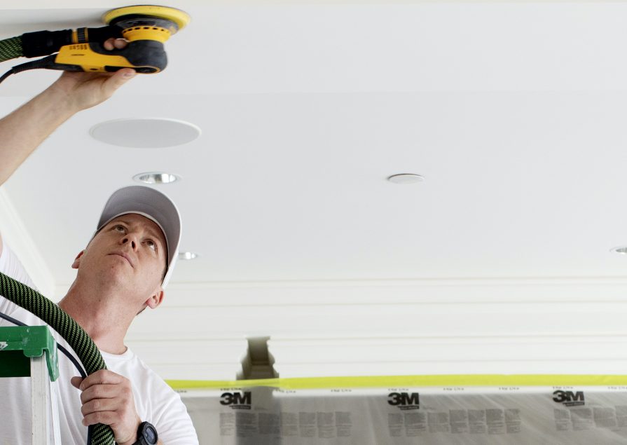 A man standing on a ladder uses a zero-dust sander on a white ceiling
